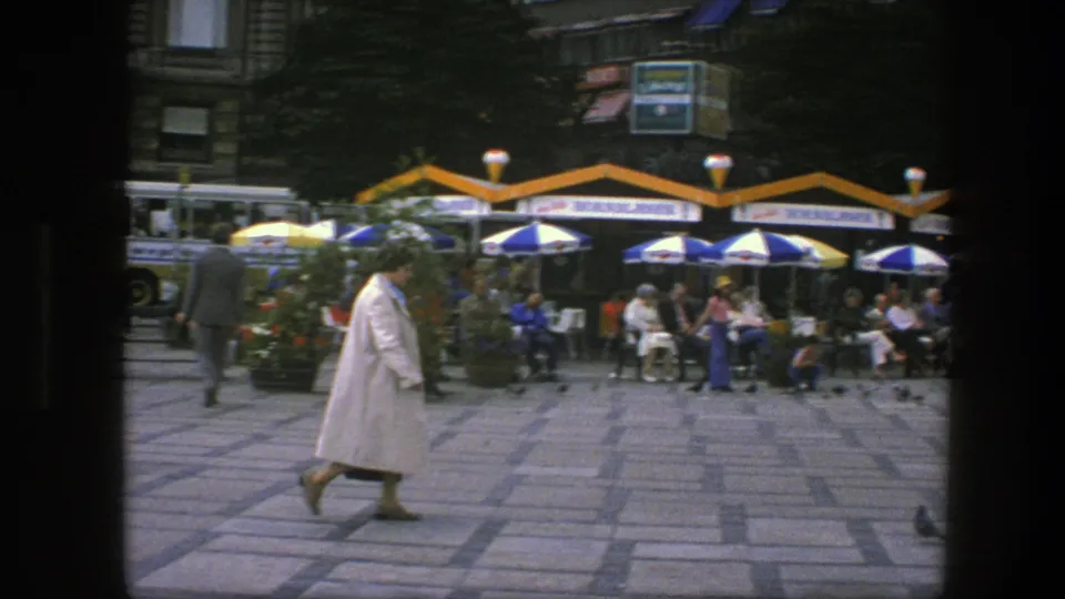 1975 Copenhagen street scene with a woman in a white coat crossing a square near cafe seating, striped umbrellas, and pigeons.