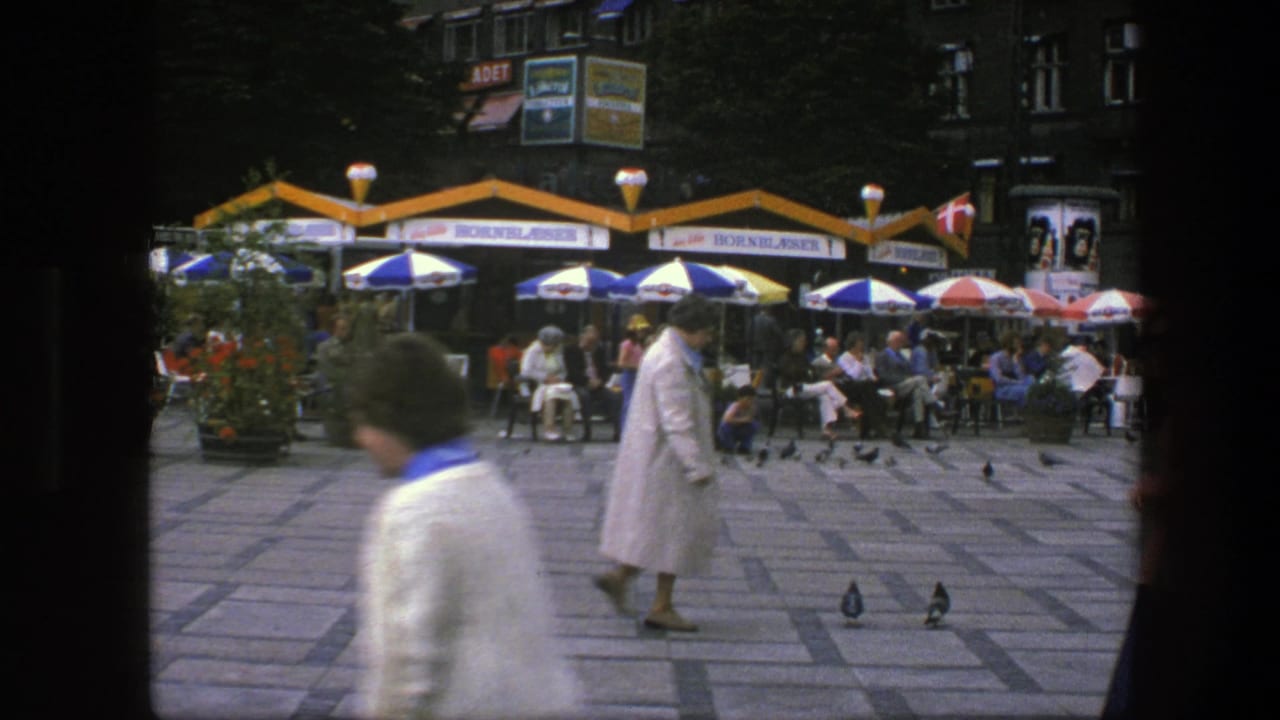 1975 Copenhagen square with a blurred foreground pedestrian, a woman in a white coat, cafe patrons, umbrellas, and pigeons.
