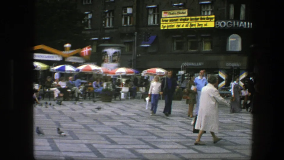 1975 Copenhagen square with the woman in white at frame edge, pedestrians, cafe umbrellas, storefront signage, and pigeons on the pavement.