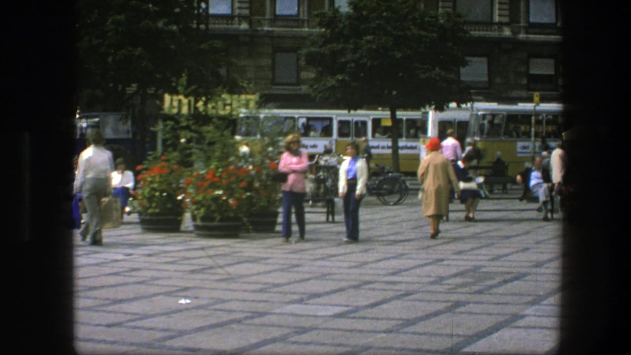 Two women walk across a paved public square with flower planters, seated people, bicycles, and buses behind them.