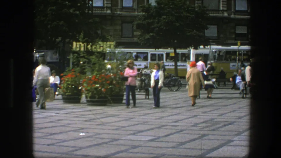 In 1975 Copenhagen, two women cross a pedestrian plaza while people sit near planters and vintage buses pass behind them.
