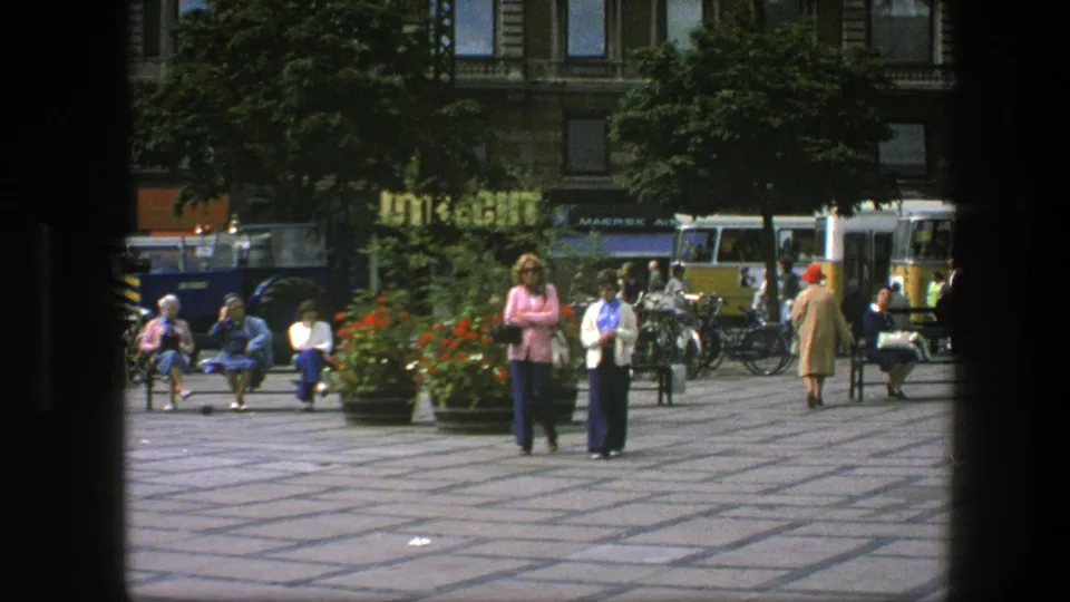 Two women in 1970s attire walk toward the camera in Copenhagen, with seated pedestrians, planters, bicycles, and buses behind them.