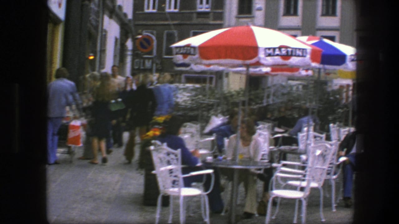 Outdoor café with striped umbrellas, white chairs, seated patrons, and pedestrians on a city sidewalk.