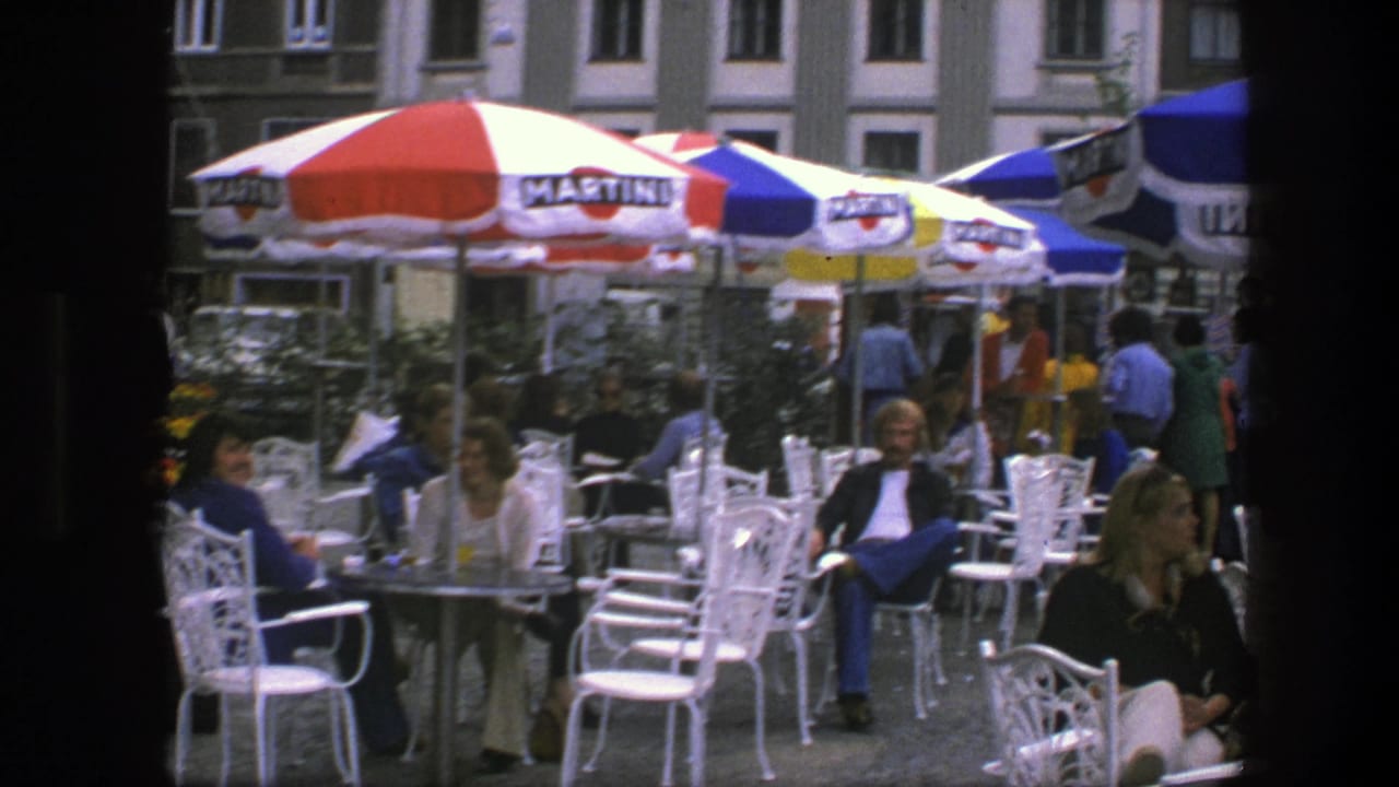 Seated café patrons under red, white, and blue umbrellas with pedestrians and buildings behind them.