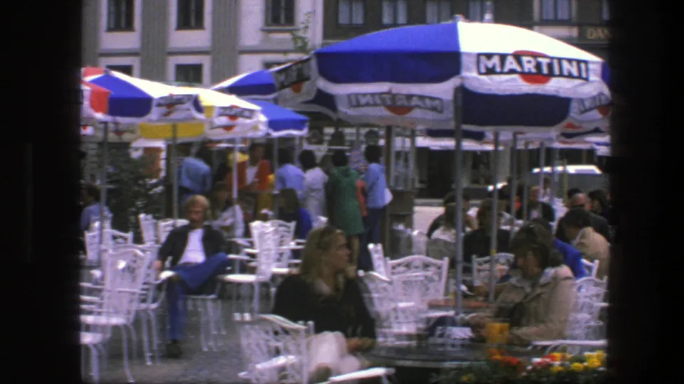 Copenhagen sidewalk café scene with seated patrons, flower planters, white chairs, and striped Martini umbrellas in 1975 home-movie footage.