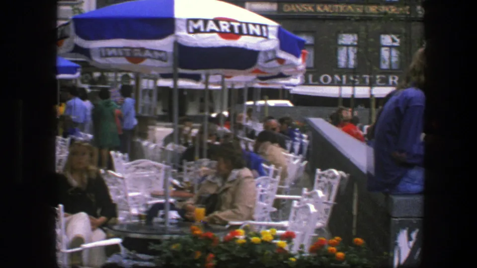 1975 Copenhagen café view with patrons at outdoor tables, Martini umbrellas, flower boxes, and storefront façades behind the seating area.