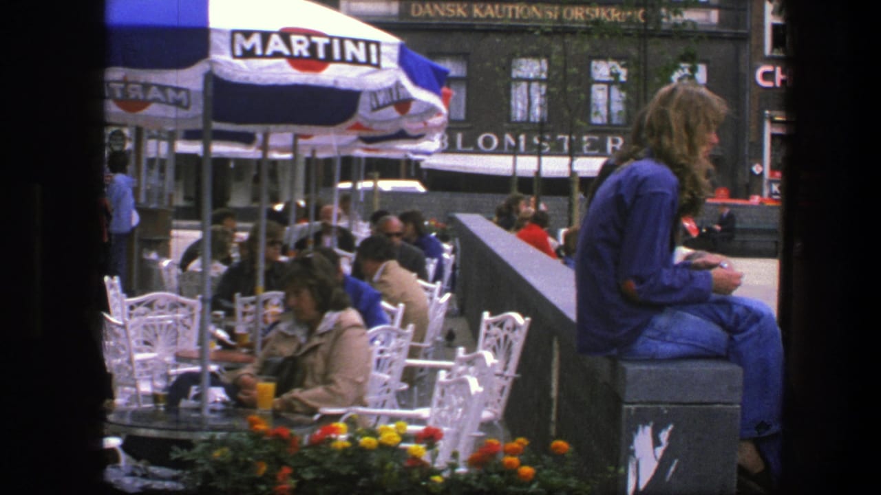 Outdoor cafe tables under Martini umbrellas with pedestrians and a seated person in blue near a city square.