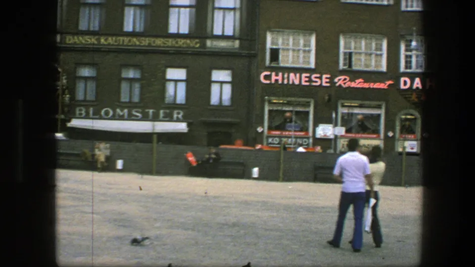 1975 Copenhagen frame with two people in the square, pigeons on the pavement, benches, Blomster lettering, and Chinese restaurant signage.