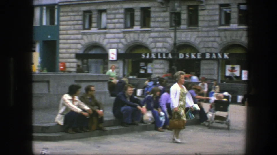 1975 Copenhagen frame with people sitting at the fountain, a woman carrying a handbag, stone buildings, and visible bank signage.