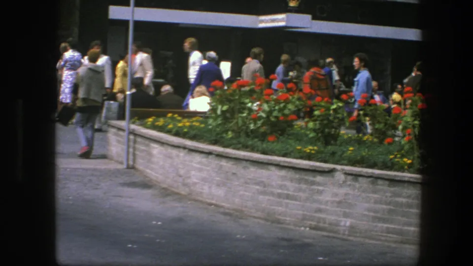 Copenhagen public-square view with pedestrians moving past a planted flower bed and storefront area in 1970s home-movie footage.