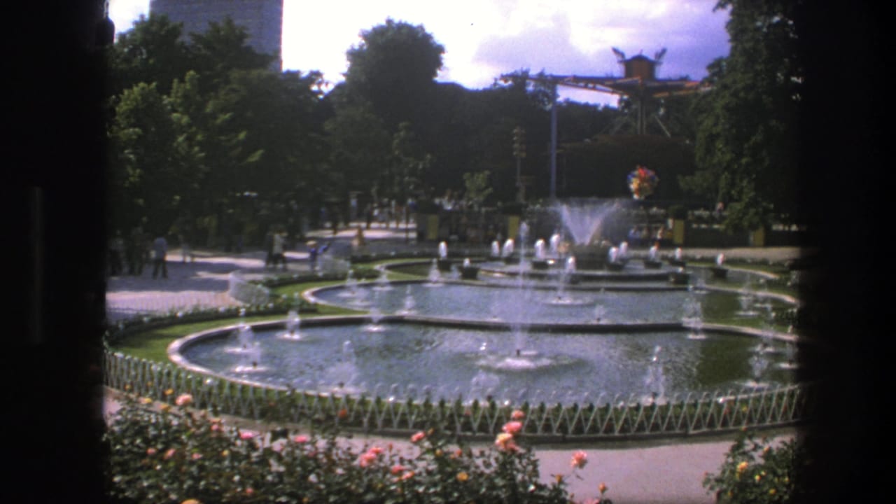 Circular park fountains spraying water with flowers, trees, paths, and distant visitors.