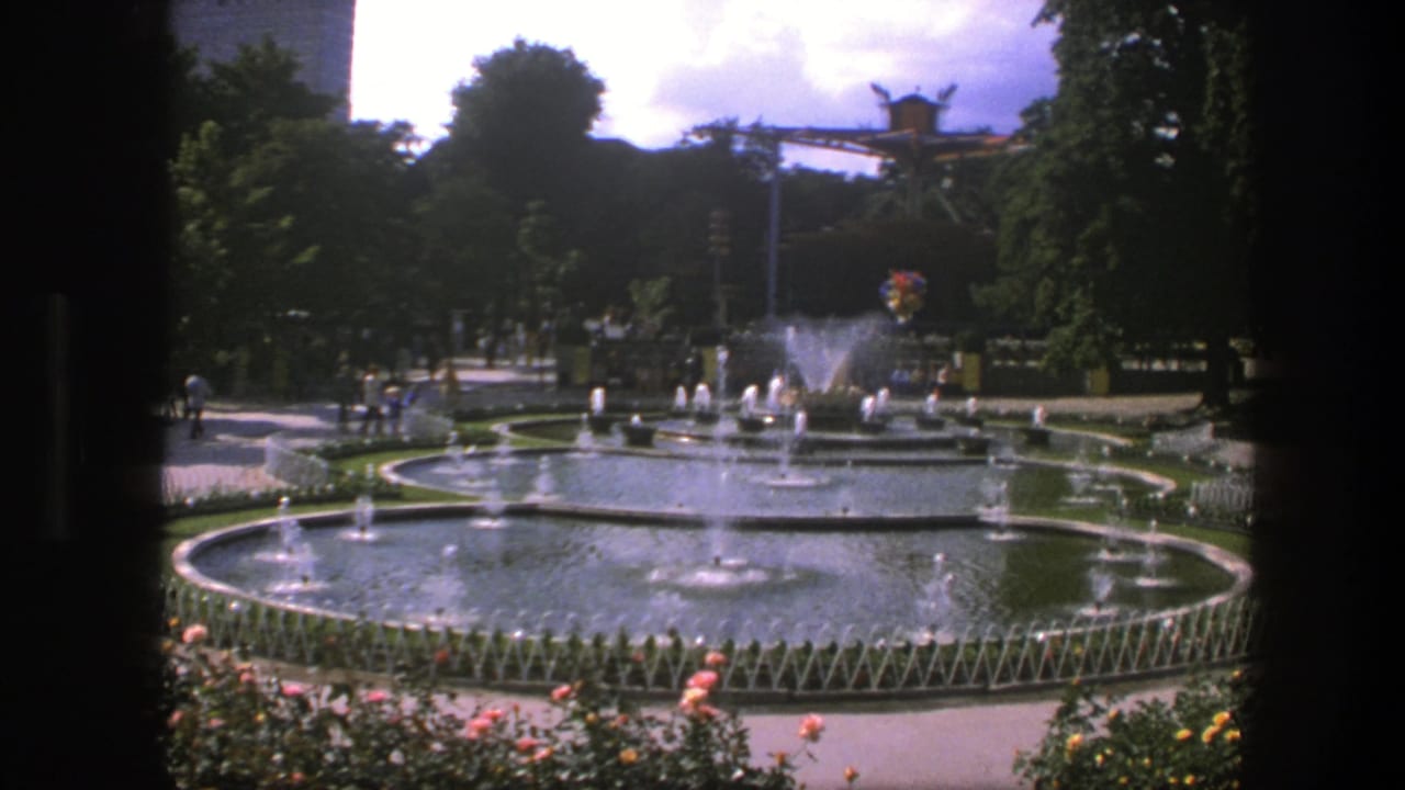 1975 Copenhagen fountain scene with multiple circular basins, spraying jets, garden planting, shaded paths, and distant park visitors.