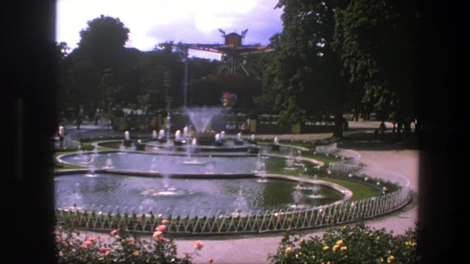 1975 Copenhagen park scene showing circular fountains, water spray, flower beds, low fencing, trees, walkways, and distant visitors.