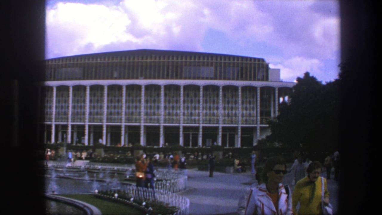 Pedestrians walk near a modernist building with a curved facade and patterned glass.