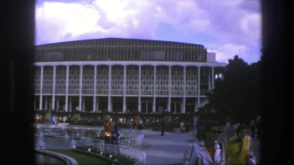 1975 Copenhagen home-movie frame showing visitors near a modernist public building, fountain area, trees, and garden planting.