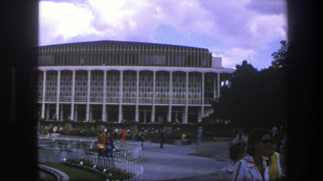 Visitors gather and walk in front of a large modernist building and garden path.