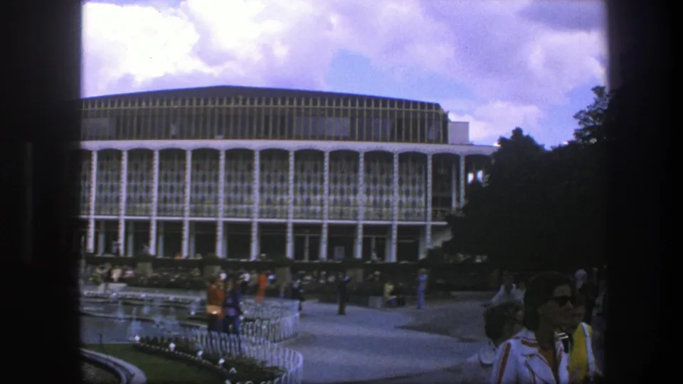 1975 Copenhagen frame with people in 1970s clothing crossing a public square before a modernist facade and fountain landscaping.