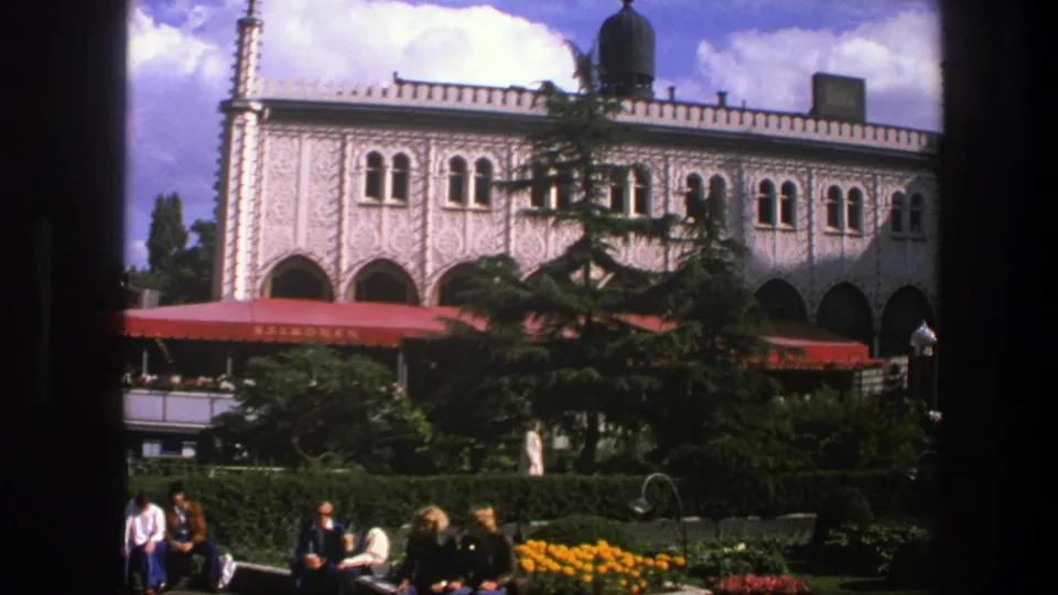 1975 Copenhagen frame showing an ornate arched exterior with red awnings, seated visitors, hedges, flowers, and trees.