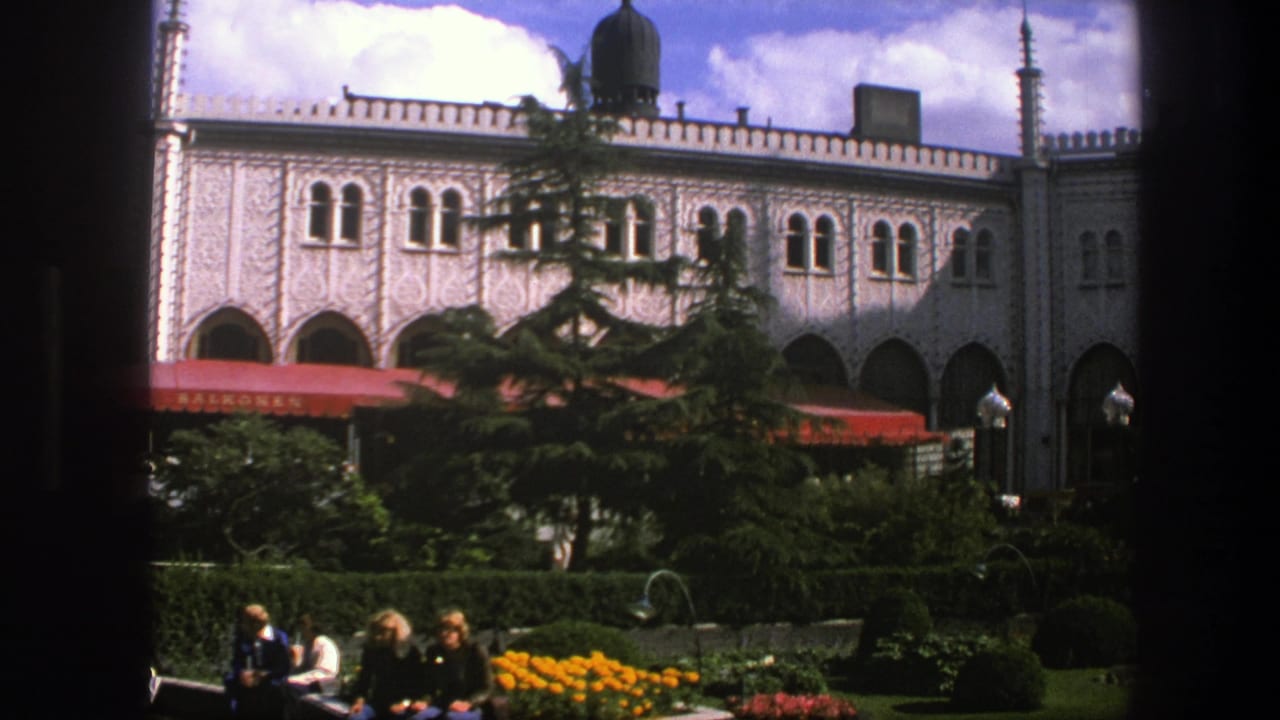 1975 Copenhagen home-movie view of a formal garden with visitors, yellow flowers, trees, red awnings, and arched windows.