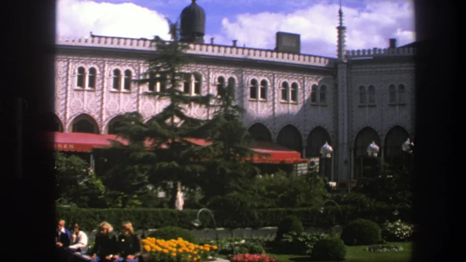 1975 Copenhagen 8mm still with seated garden visitors, yellow flowers, hedges, red awnings, and a pale ornate building facade.