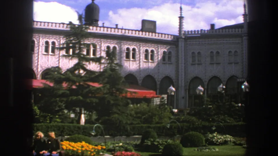 1975 Copenhagen home-movie frame showing garden plantings, seated visitors, red awnings, arched openings, and pale decorative architecture.