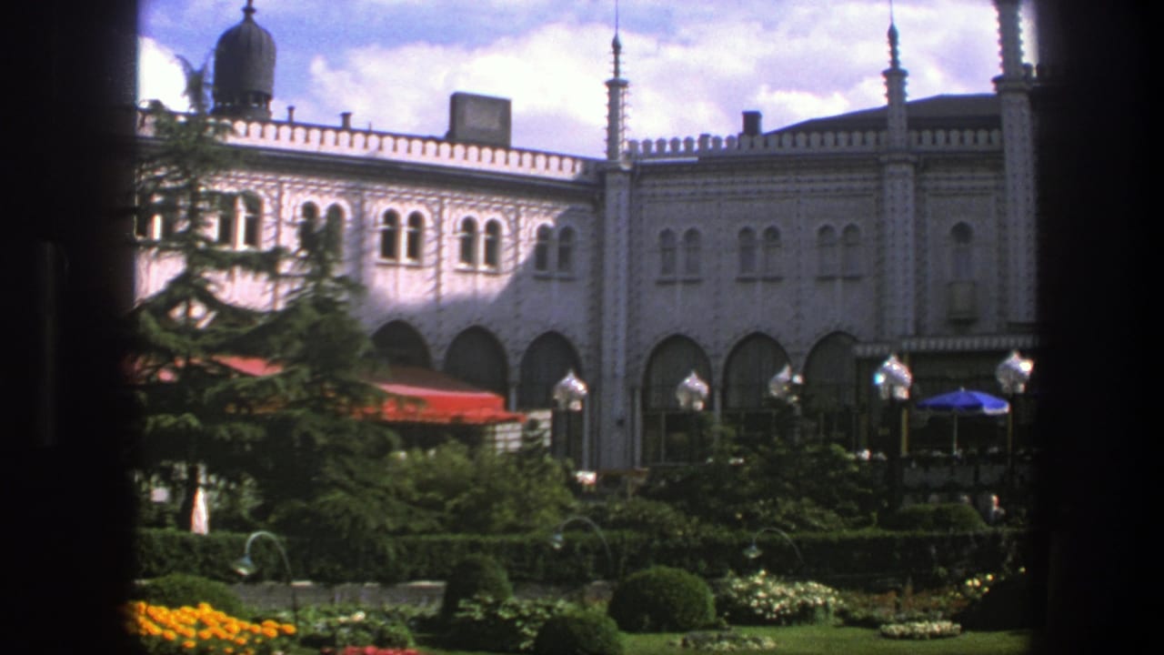 1975 Copenhagen frame with arched windows, pale decorative facade, planted garden, lamp posts, and cafe umbrellas.
