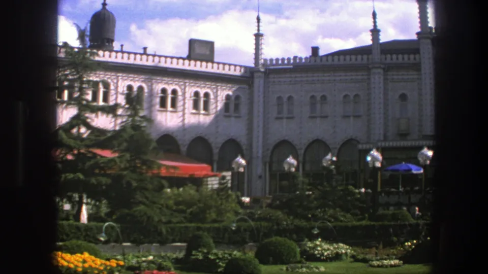 1975 Copenhagen frame with arched windows, pale decorative facade, planted garden, lamp posts, and cafe umbrellas.
