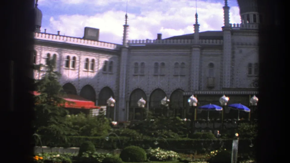 1975 Copenhagen home-movie frame showing the ornate building, clipped garden, red and blue umbrellas, and small distant visitors.