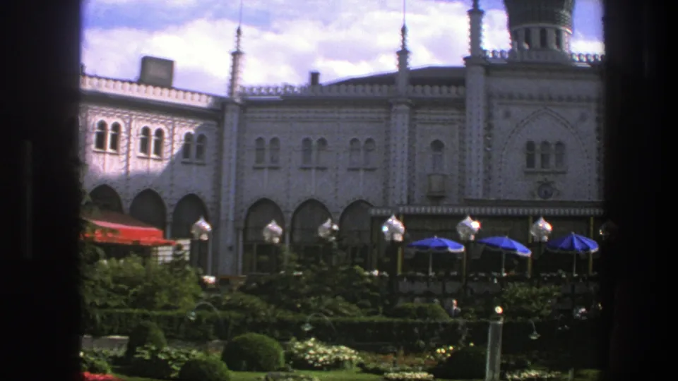 1975 Copenhagen frame with ornate arches, spires, garden planting, lamp posts, blue umbrellas, and distant people.