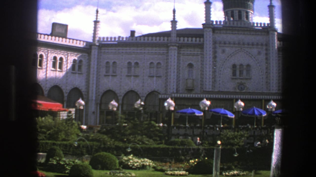 Ornate building facade seen across a garden with blue umbrellas and dark foreground posts.