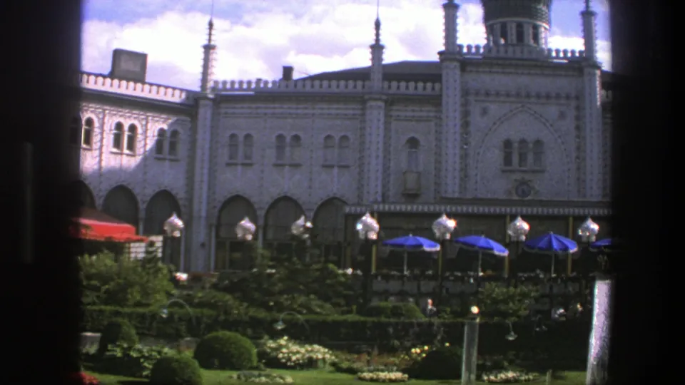 1975 Copenhagen 8mm frame showing an ornate Gothic Revival-style facade beyond hedges, flowers, terrace umbrellas, and small visitors.