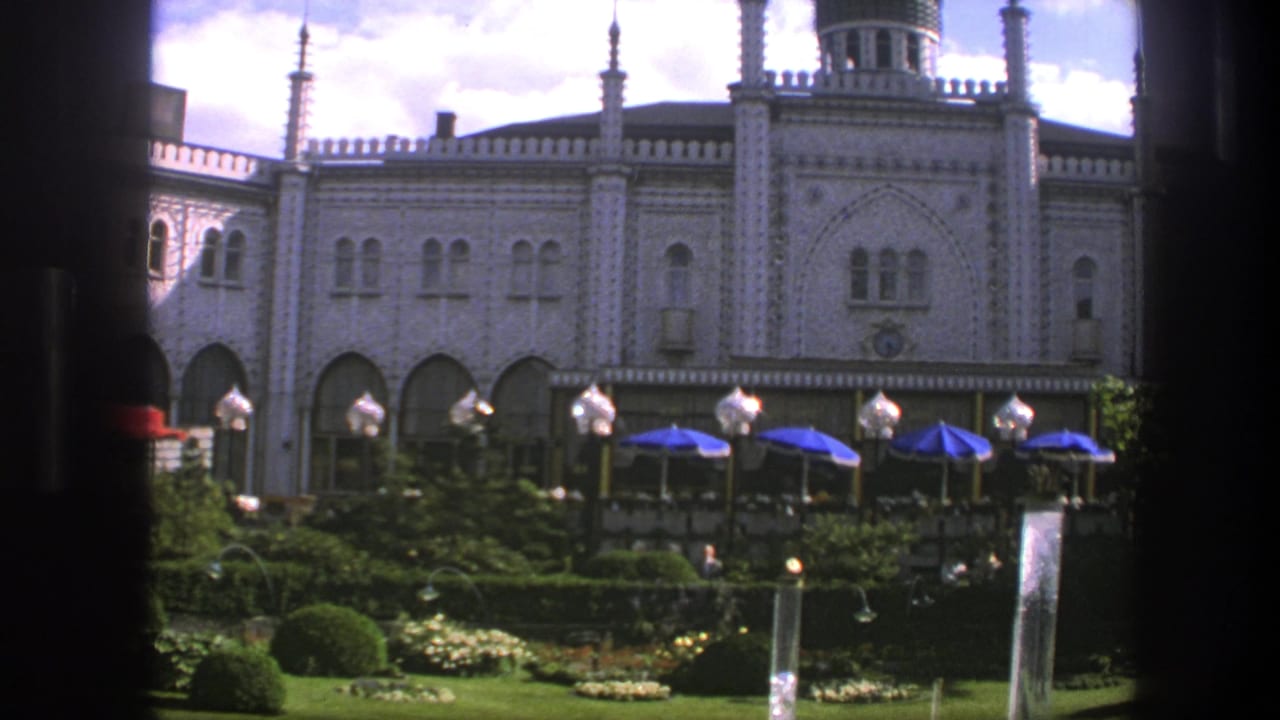 1975 Copenhagen frame with a decorative facade, arched lower level, blue terrace umbrellas, globe lamps, and greenery in front.