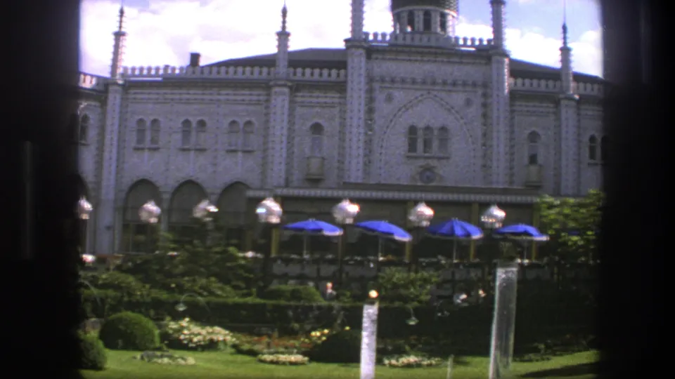 Soft 1975 Copenhagen 8mm view of an ornate facade framed by dark posts, with hedges, flowers, lamps, and blue umbrellas below.