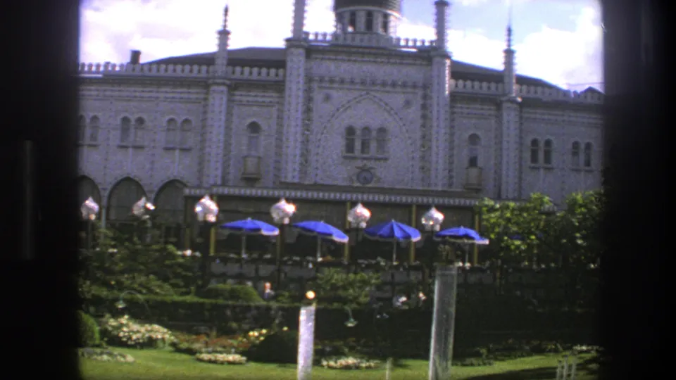 1975 Copenhagen 8mm frame showing the ornate building front, garden plantings, blue umbrellas, globe lamps, and small distant visitors.