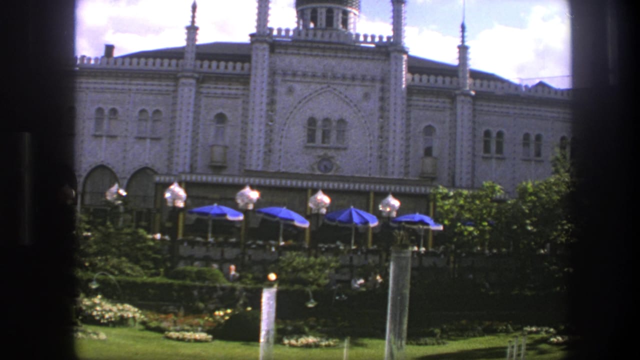 Ornate palace facade behind gardens, blue umbrellas, lamps, and a fountain, partly framed by dark window edges.