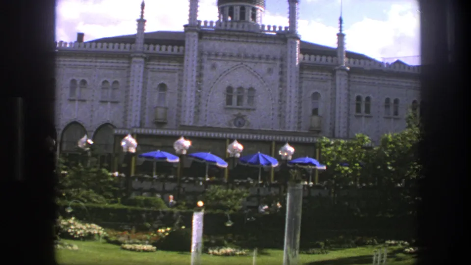 1975 Copenhagen home-movie view of a palace garden with blue terrace umbrellas, lamps, fountain jets, and dark foreground window framing.