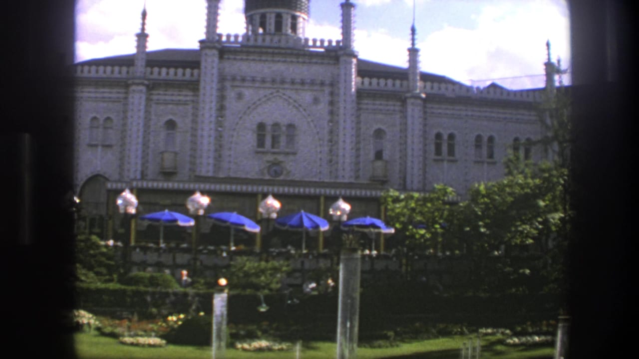 Palace exterior with tower, arched windows, garden trees, blue umbrellas, lamps, and fountain spray.