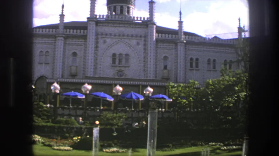 Copenhagen palace exterior in 1975 with a manicured garden, terrace umbrellas, decorative lamps, and fountain water under a bright sky.