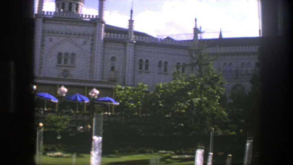 1975 Copenhagen garden view with the palace facade partly off center, blue umbrellas and lamps below, and fountain columns in front.