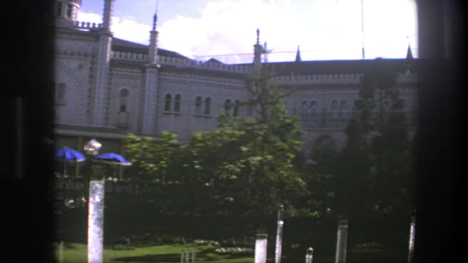 Late-frame 1975 Copenhagen view of the palace garden, partly obscured by dark window edges, with trees, blue umbrellas, lamps, and fountain water.