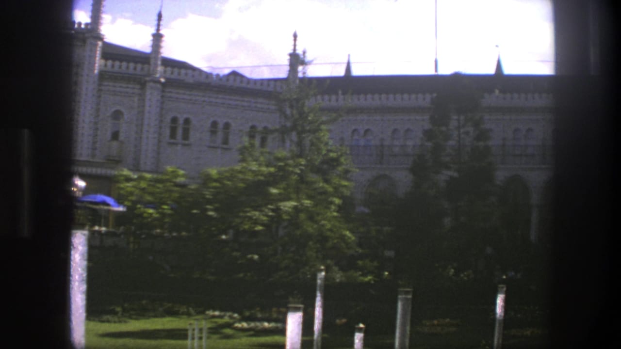 Ornate pale building seen through dark window framing with trees and garden posts in front.
