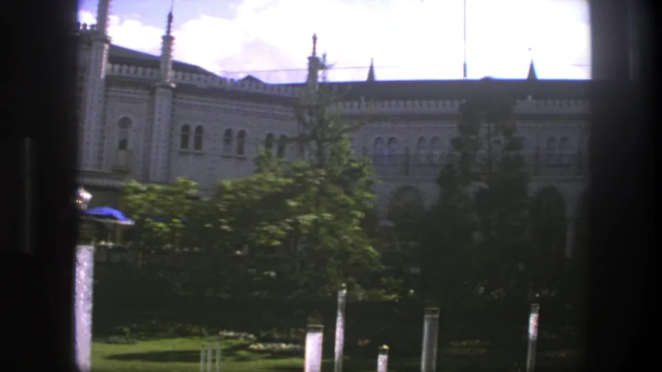 1975 Copenhagen home-movie view of an ornate building facade seen through a window, with trees, lawn, posts, and a small blue umbrella.