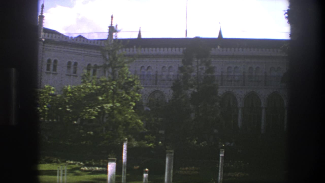 Window-framed view of a decorative building facade with trees and garden foreground.