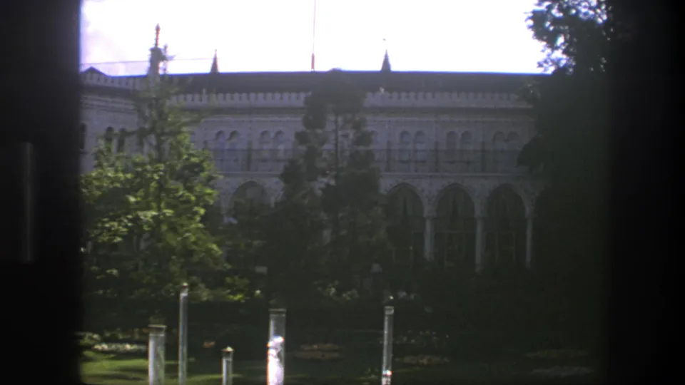 1975 Copenhagen home-movie frame with a closer view of arched windows and columns behind trees, filmed through dark vertical window framing.
