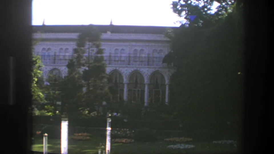 1975 Copenhagen 8mm view of a pale ornate building, arched windows, trees, lawn, and garden posts with dark window framing at the side.
