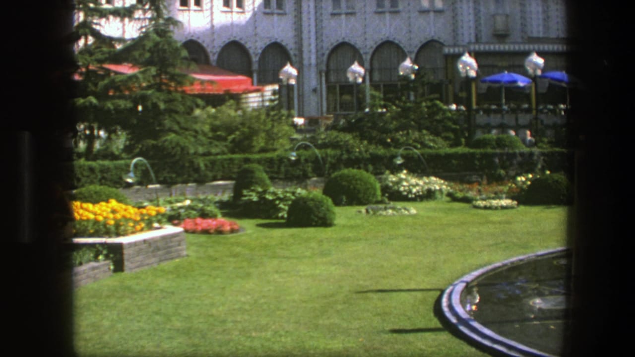 1975 Copenhagen garden view with clipped lawn, yellow and red flowerbeds, shrubs, and an arched building behind cafe umbrellas.