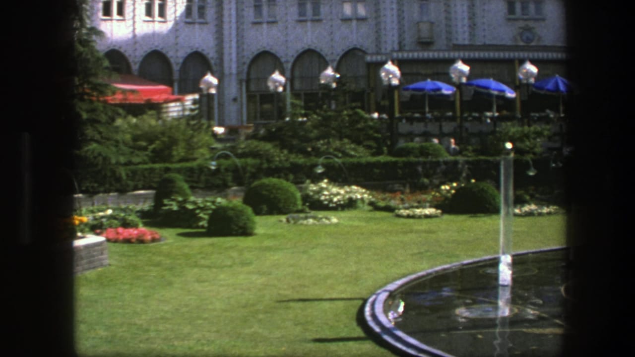 1975 Copenhagen garden scene with a curved fountain pool, vertical water jet, trimmed lawn, shrubs, and blue umbrellas near the building.