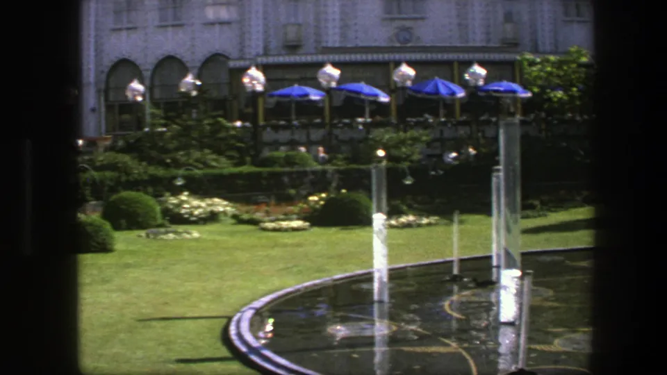 1975 Copenhagen fountain view with multiple water jets, reflective pool, manicured lawn, flowerbeds, shrubs, and distant cafe seating.