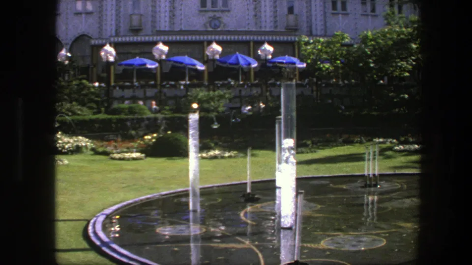 1975 Copenhagen garden and fountain with bright water jets, formal plantings, blue umbrellas, and distant visitors near an arched facade.