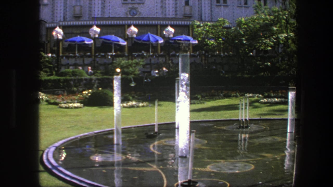 1975 Copenhagen 8mm frame showing fountain jets in a reflecting pool, green lawn, blue umbrellas, trees, and an ornate building beyond.